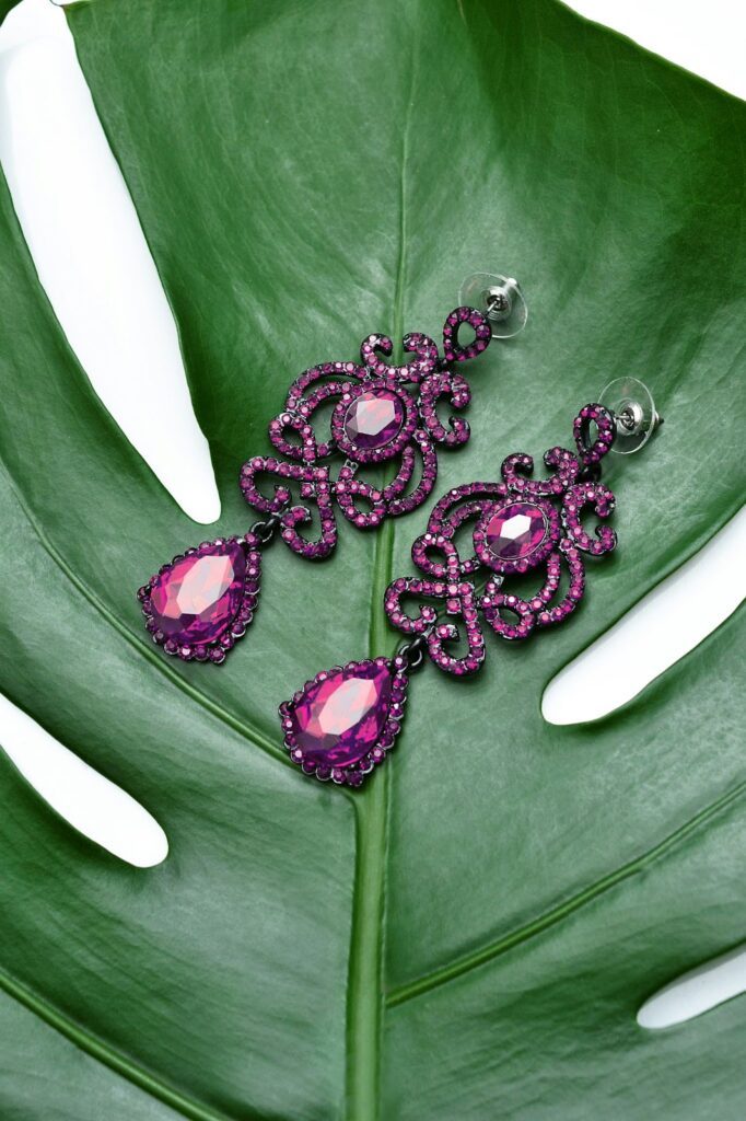 Intricate purple gemstone earrings with a bold, ornate design, featuring teardrop and round-cut stones set against a lush green leaf background.