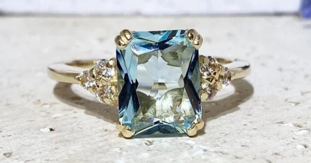 Elegant aquamarine gemstone ring with a rectangular-cut center stone, set in a gold band, flanked by clusters of sparkling diamonds on each side.