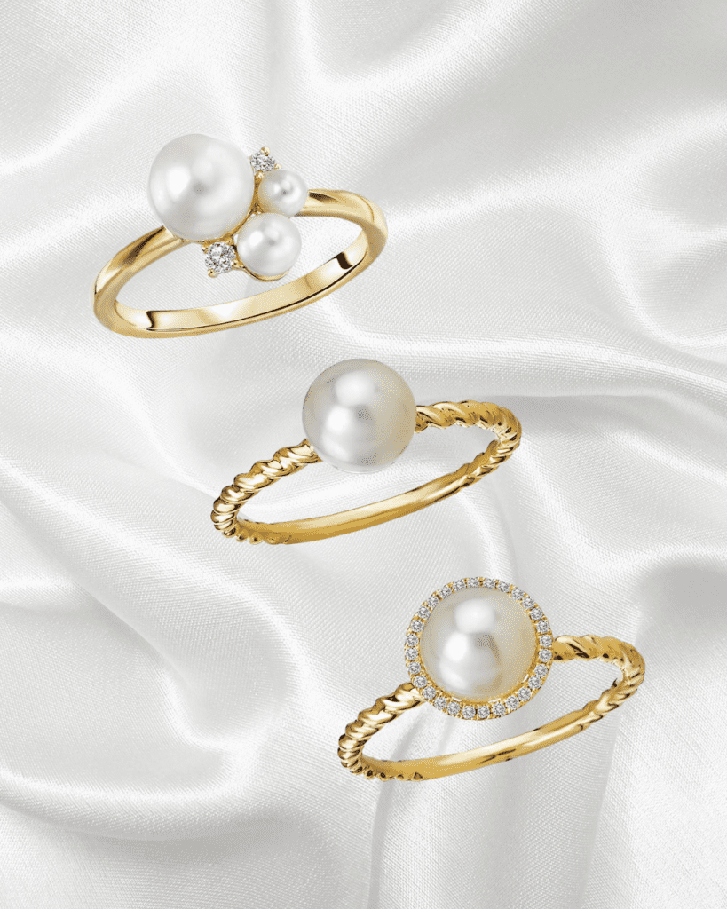 Gold ring with three elegant pearls of varying sizes accented with sparkling diamonds, set against a soft satin white background.