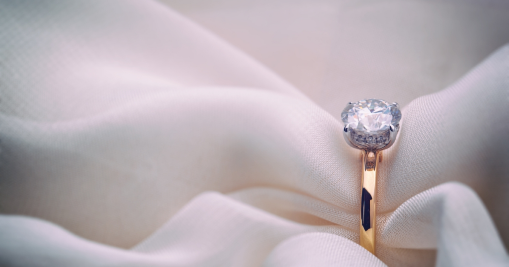 Stunning gold engagement ring with a round-cut diamond, elegantly displayed on soft, draped white fabric, highlighting the stone's brilliance.