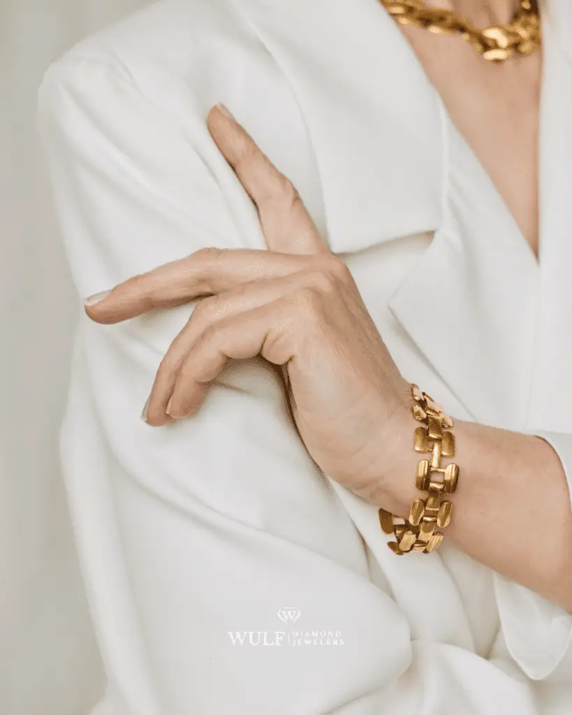 Close-up of a woman's hand wearing a gold chain bracelet, styled with a white blazer and matching gold necklace, showcasing elegant fashion.