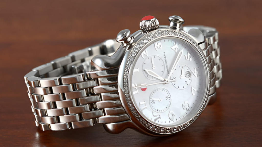 Close-up of a silver chronograph watch with a mother-of-pearl dial, diamond-accented bezel, and red crown, set against a wooden background.