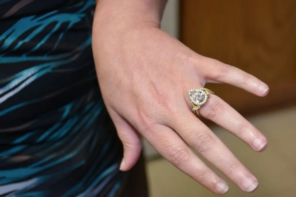 A close-up of a hand wearing a pear-shaped diamond engagement ring with a gold band and smaller diamonds around the stone. The hand is resting on a surface.