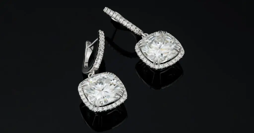 A pair of diamond drop earrings with a cushion-cut center stone surrounded by smaller diamonds in a halo setting. The earrings have a white gold hook closure.