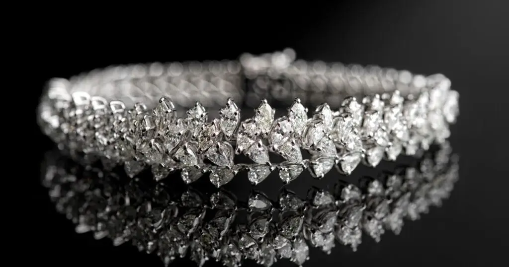 A diamond tennis bracelet with a row of pear-shaped diamonds set in a white gold band. The bracelet reflects on a black surface.