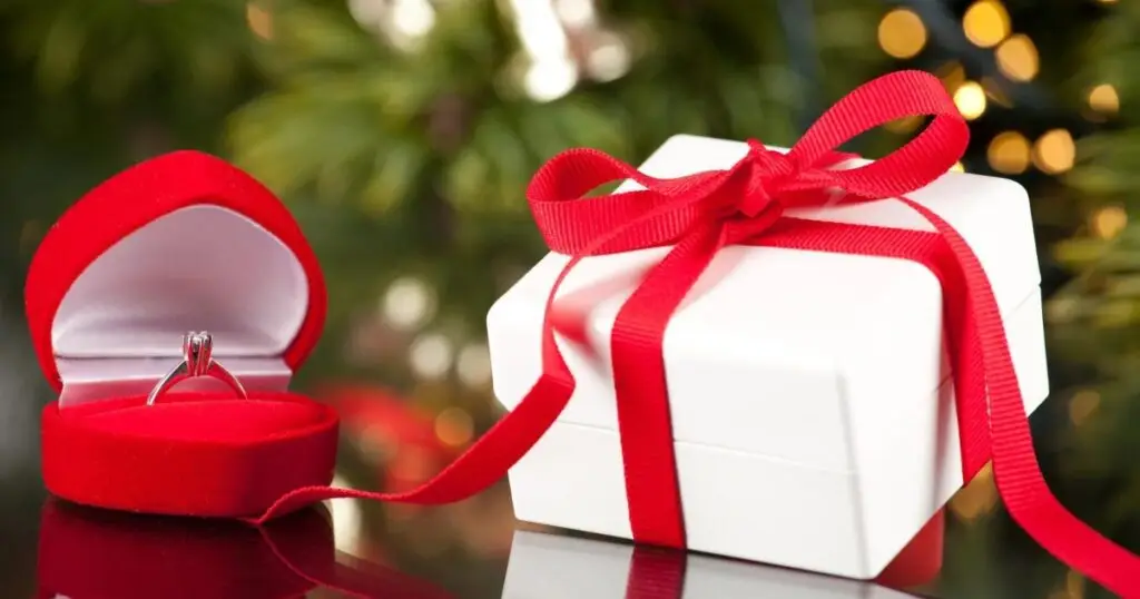 A red gift box open on a green background, revealing a diamond engagement ring. A Christmas wreath and ornaments decorate the scene.