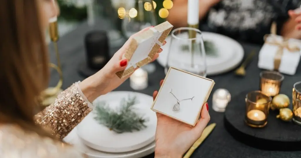 A woman holding an open jewelry box with a diamond pendant necklace, preparing to present it as a gift at a candlelit dinner table.