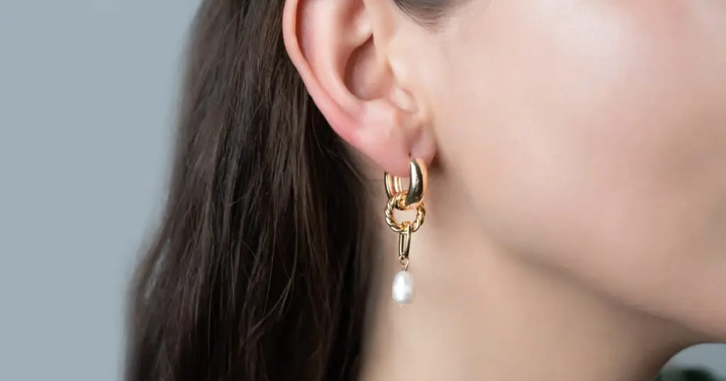 Close-up of a woman's ear wearing a stylish gold hoop earring with a twisted chain link and a dangling pearl, set against a light background.