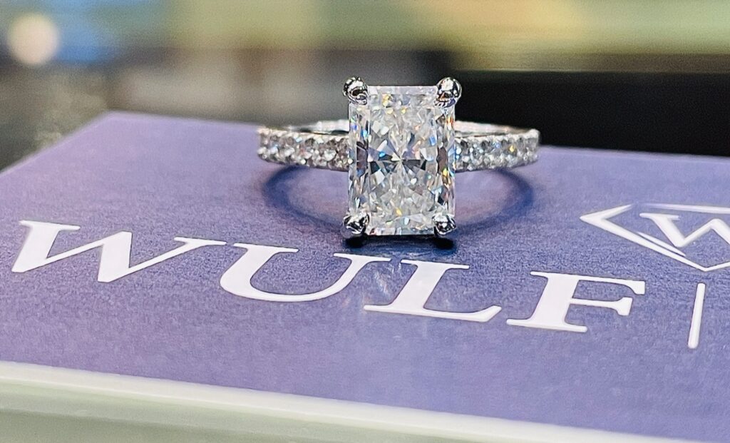 Radiant-cut diamond engagement ring with a shimmering band of accent diamonds, elegantly displayed on a purple WULF-branded background.