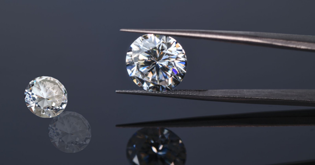 A large, round diamond held in tweezers with a smaller diamond resting on a black surface.