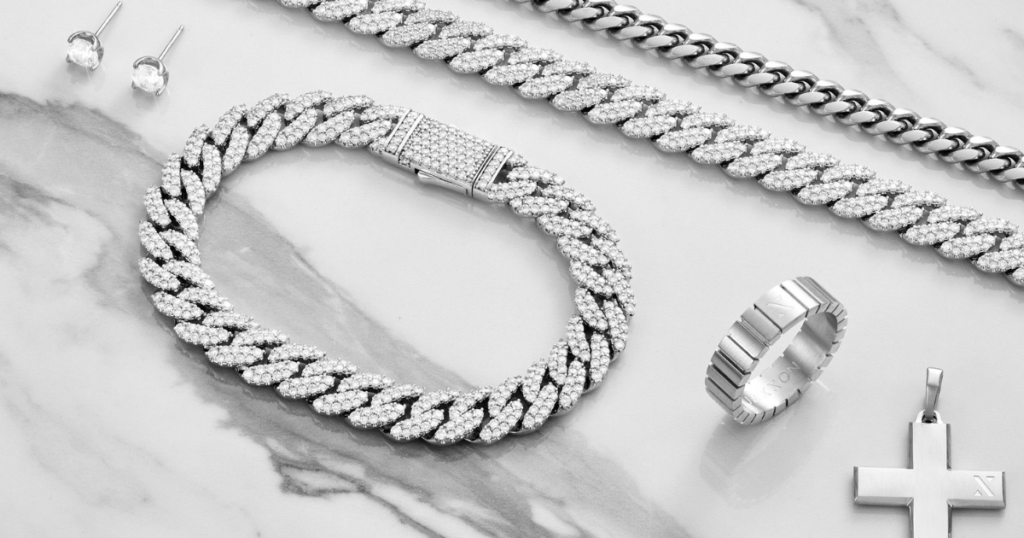 A collection of iced-out jewelry pieces, including a Cuban link chain, a ring, a cross pendant, and stud earrings, displayed on a marble background.