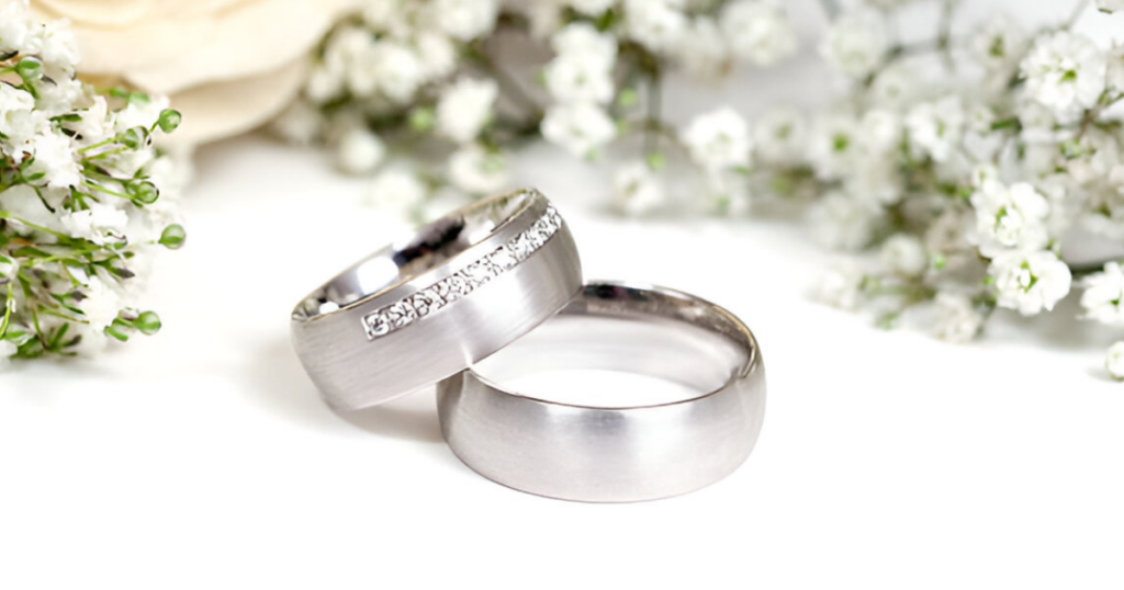 Two wedding bands, one with a row of diamonds, resting on a white surface with white flowers.