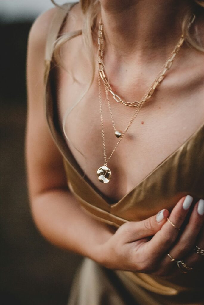 Close-up of a woman's layered gold necklaces with textured pendants, complemented by her gold rings and white manicured nails, adding elegance.