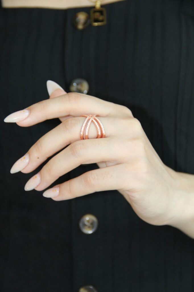 Elegant hand with long, almond-shaped nails holding a rose gold statement ring with intricate design, set against a black outfit with button details.