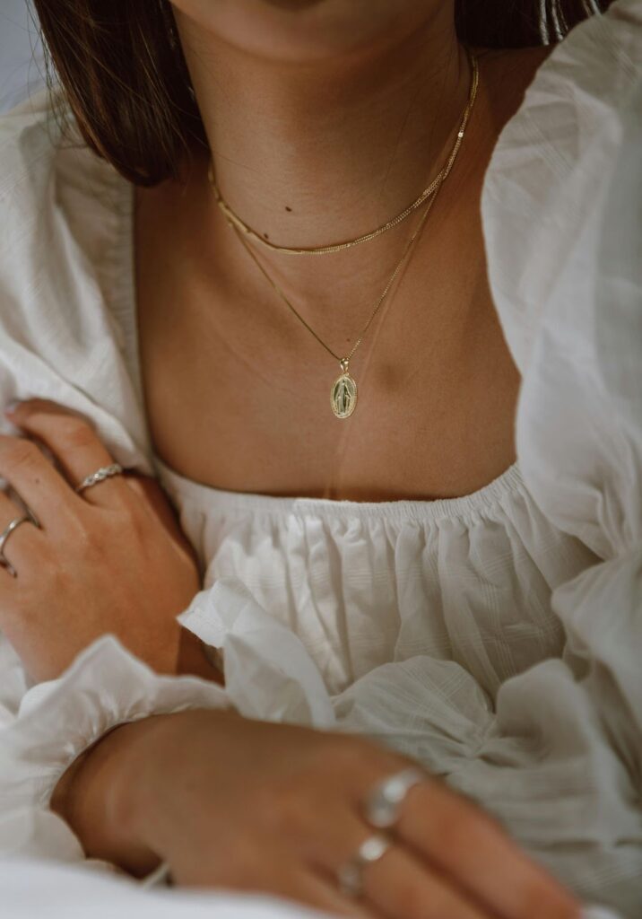 A close-up of a person’s collarbone showcasing layered gold necklaces and silver rings, set against a soft, white textured top.