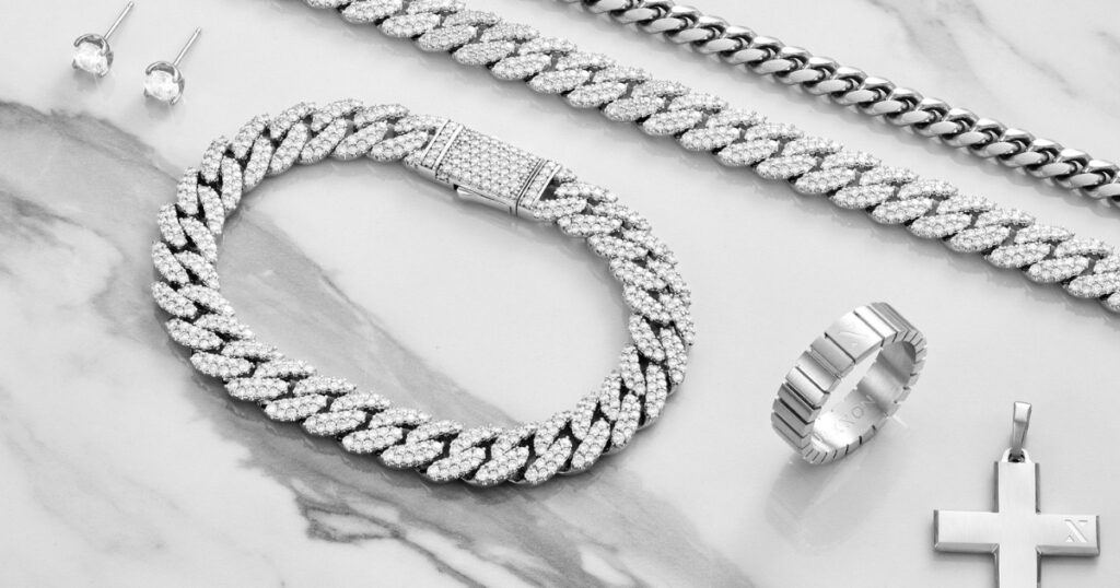 A collection of iced-out jewelry pieces, including a Cuban link chain, a ring, a cross pendant, and stud earrings, displayed on a marble background.