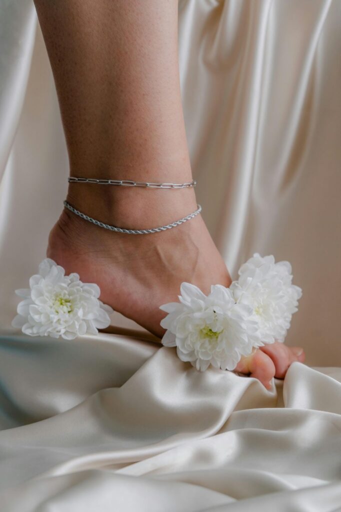 Close-up of a woman's ankle adorned with delicate silver anklets, surrounded by white flowers on a soft, cream satin fabric background.