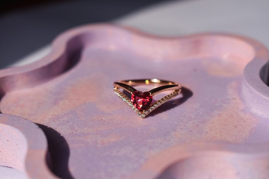 A heart-shaped ruby ring set in rose gold with diamond accents, resting on a pink dish.