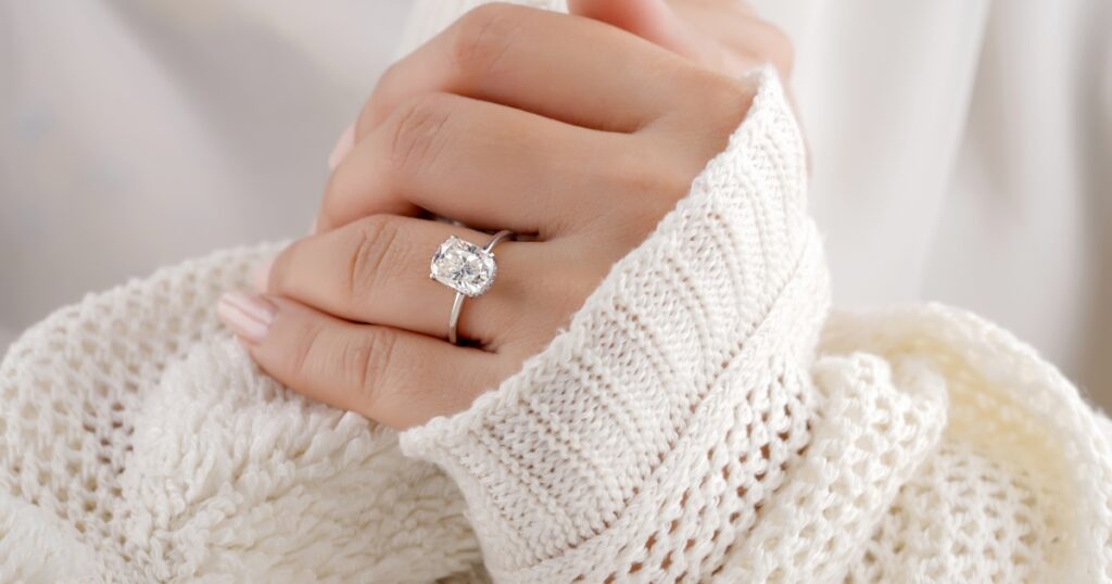 Close-up of a hand wearing a classic cushion-cut diamond solitaire ring on a simple band, paired with a cozy white knitted sweater.