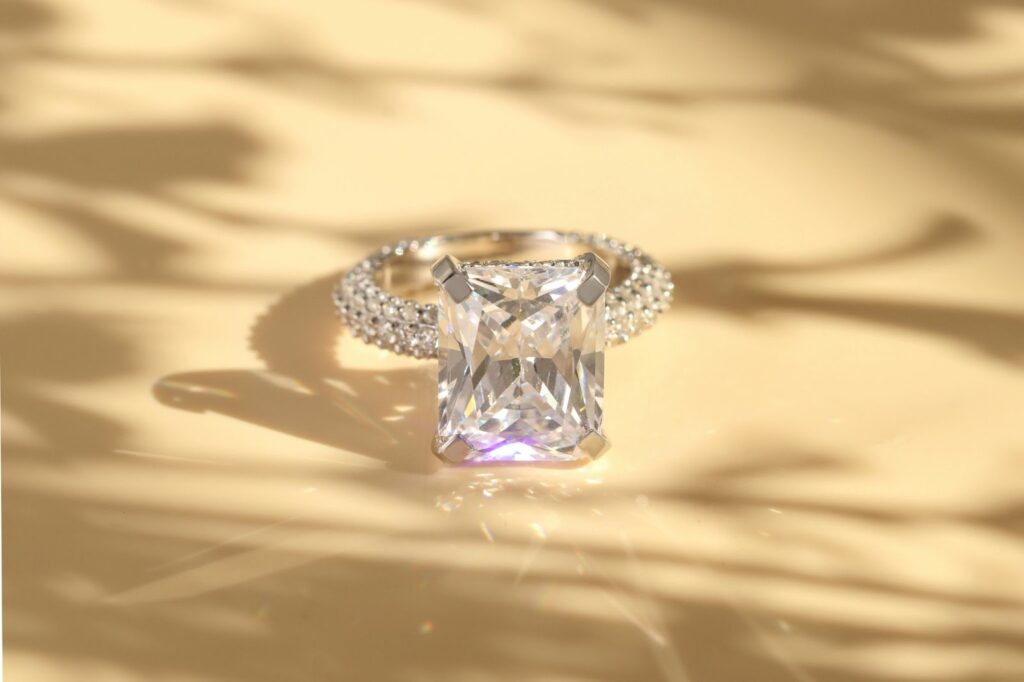 A stunning emerald-cut diamond ring with a diamond-studded band, resting on a soft, light-colored surface.