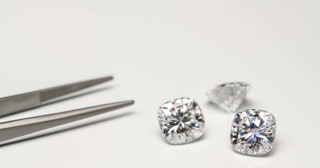 A pair of tweezers holding a cushion-cut diamond, with two more cushion-cut diamonds and a triangular diamond on a white surface.