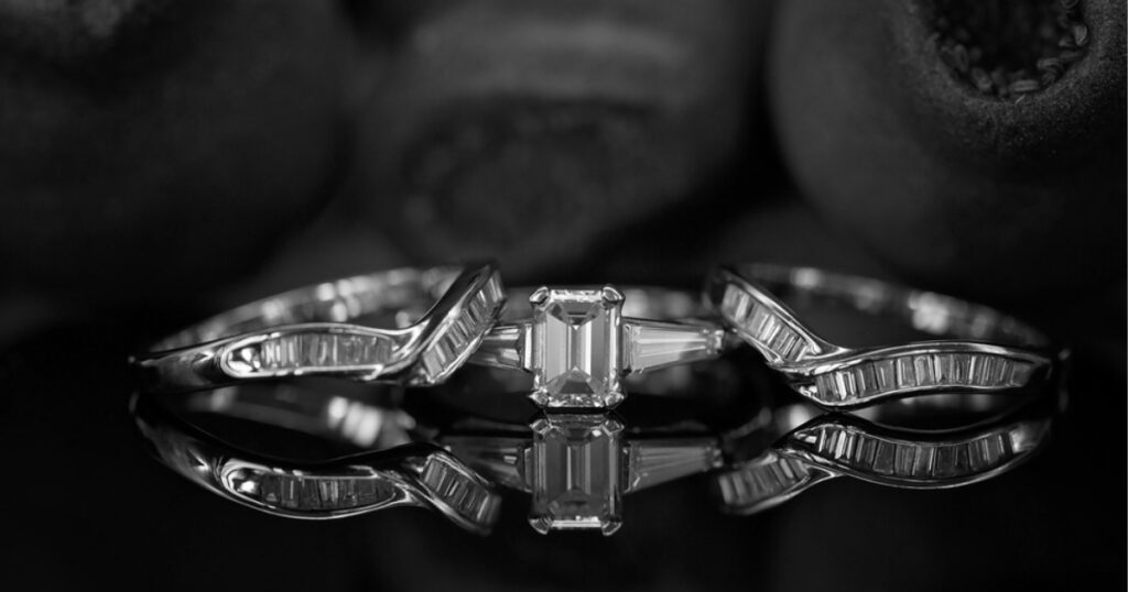 A black and white photo of a wedding band set, with an emerald-cut center stone and two smaller stones on the band, reflecting on a dark surface.