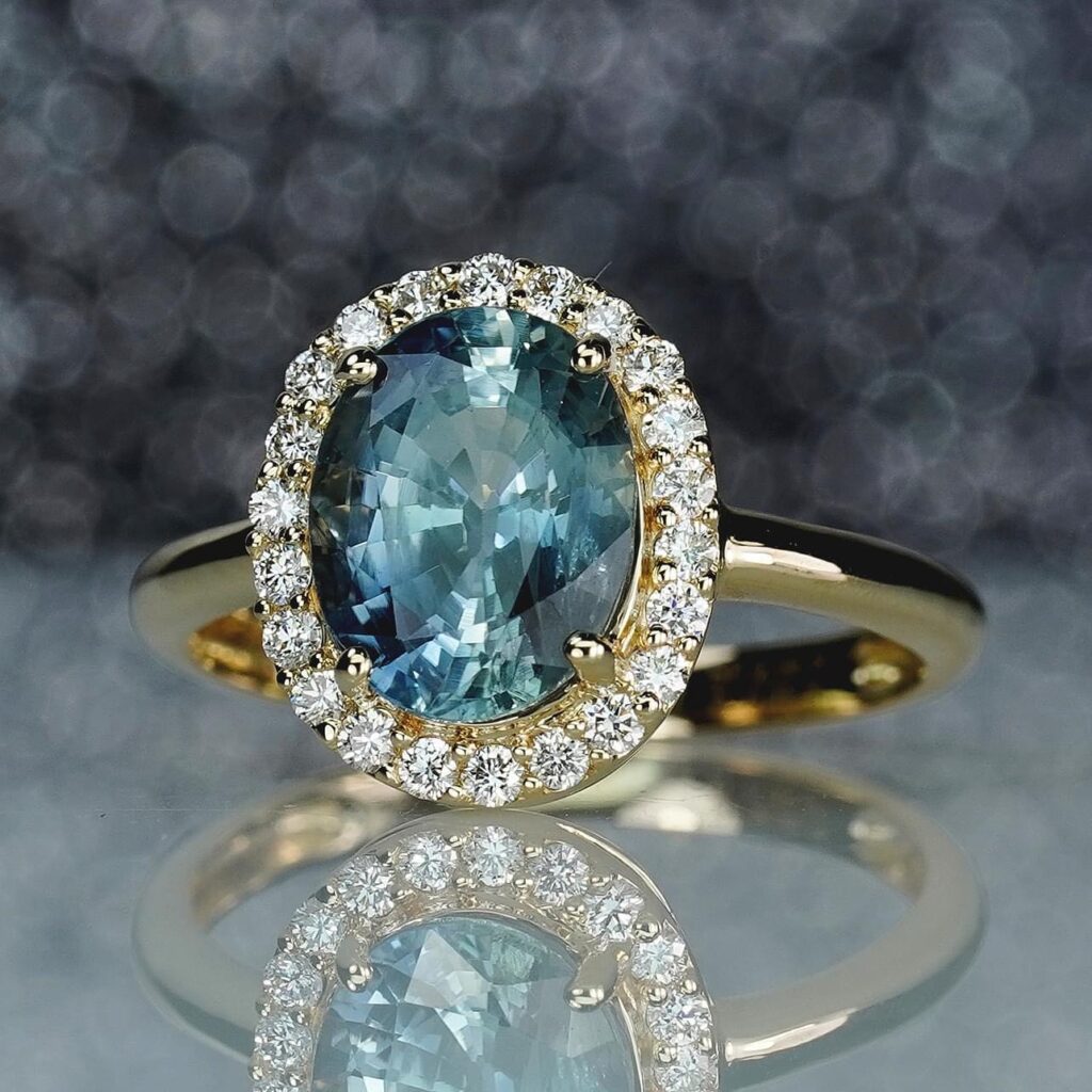 A gold ring with an oval blue sapphire center stone surrounded by a halo of diamonds, reflecting on a sparkling background.