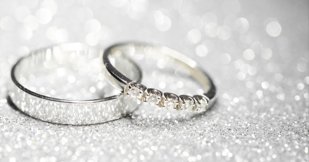 A pair of wedding rings resting on a shimmering silver background. One ring is plain, while the other has a row of diamonds.