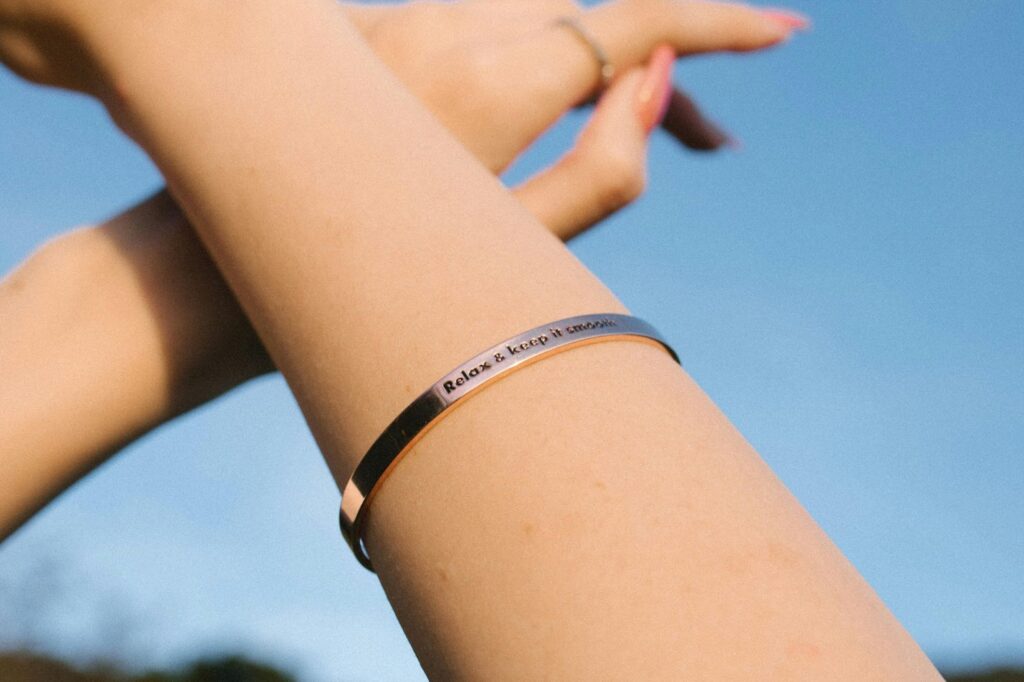 A woman's arm with a silver bracelet engraved with the words "Relax & Keep It Simple" against a blue sky background.