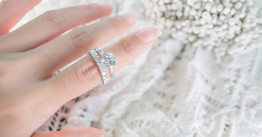 A hand wearing a diamond engagement ring and wedding band, resting on a white, lacey surface.