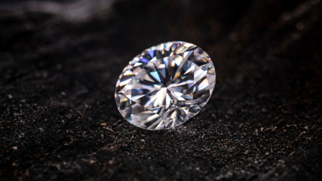 A sparkling round diamond, resting on a dark, textured surface.