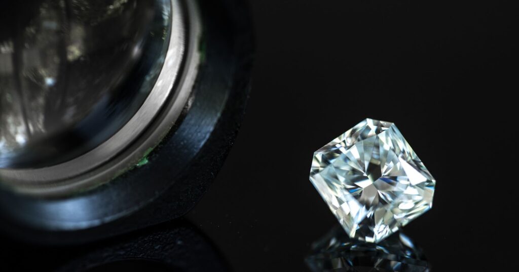 A jeweler's loupe and a sparkling radiant-cut diamond, both resting on a black surface.