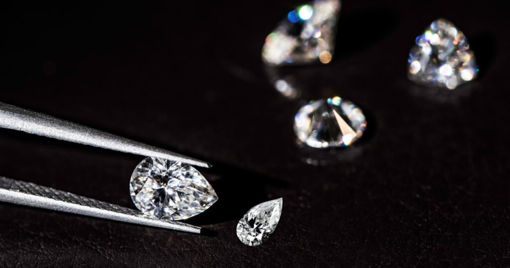 A pair of tweezers holding a pear-shaped diamond, with several other diamonds scattered on a black surface.