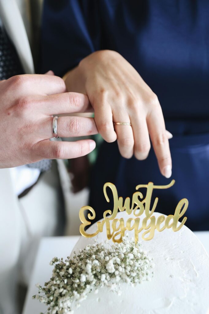 A newly engaged couple touches rings, celebrating with a "Just Engaged" cake topper.