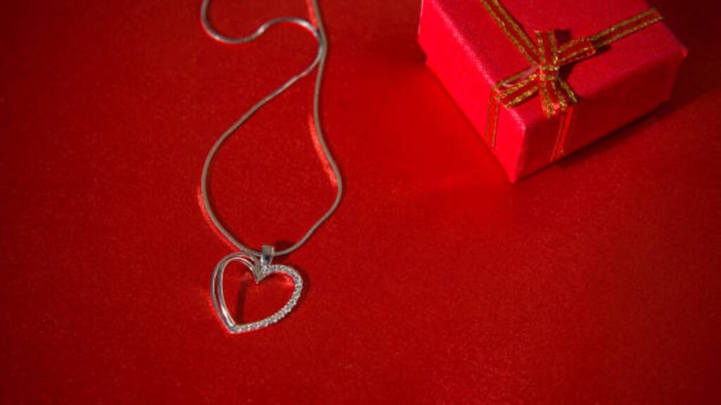 A heart-shaped necklace nestled beside a festive red gift box with a bow.