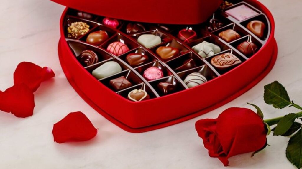 A heart-shaped box overflowing with decadent chocolates, accompanied by a single red rose and scattered petals.