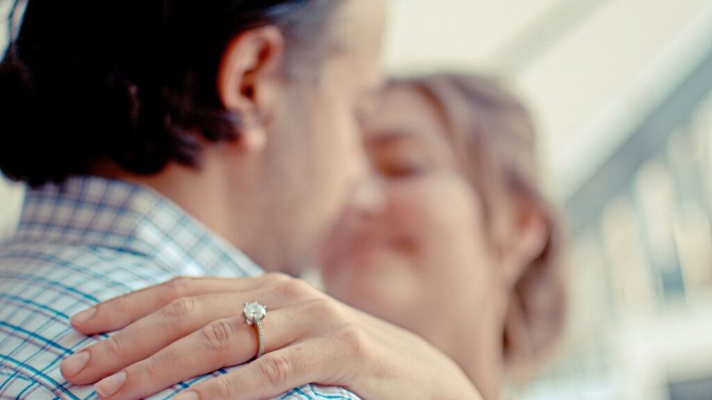 Close-up of a woman’s hand wearing a sparkling diamond engagement ring, embracing her partner in a tender, romantic moment.