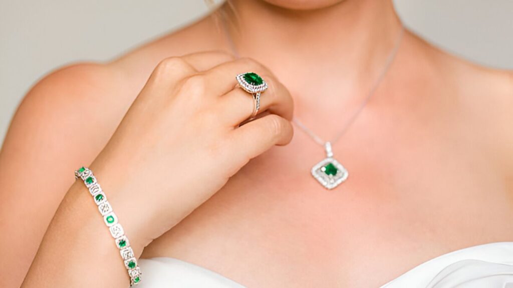 A woman's hand adjusts a ring featuring a large emerald, while wearing a matching necklace and bracelet, all set in silver.