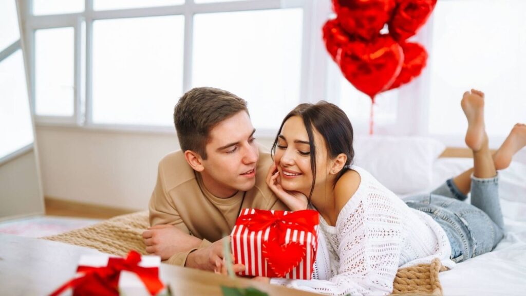 Couple sharing a romantic moment on Valentine's Day, with red heart balloons and a beautifully wrapped gift box creating a festive atmosphere.