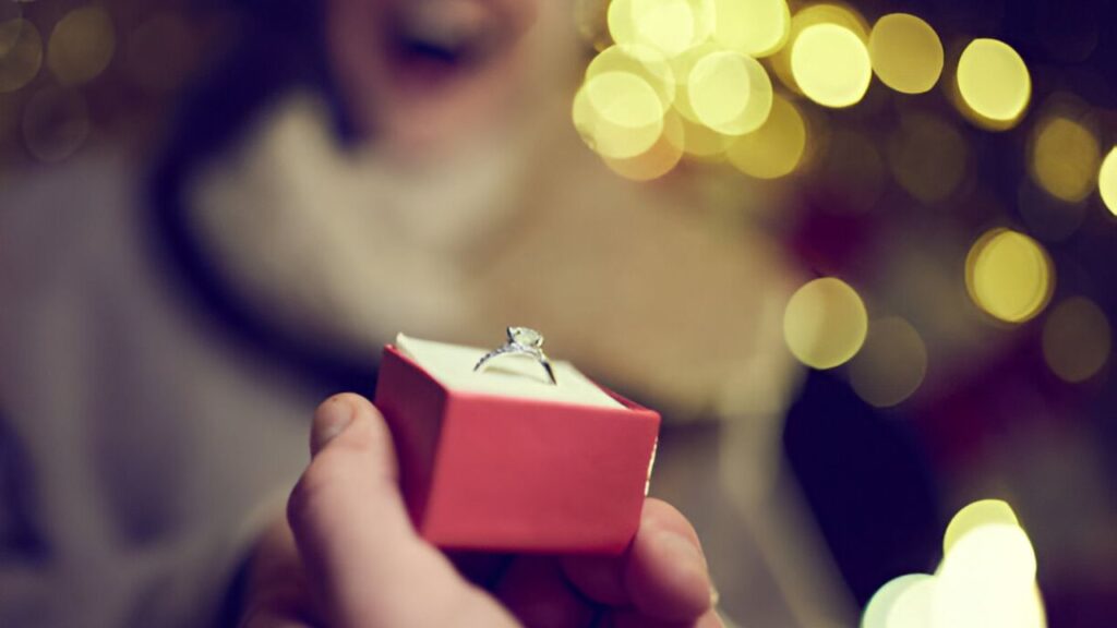 A hand holds out a red box with a sparkling diamond ring, capturing a moment of surprise and joy.