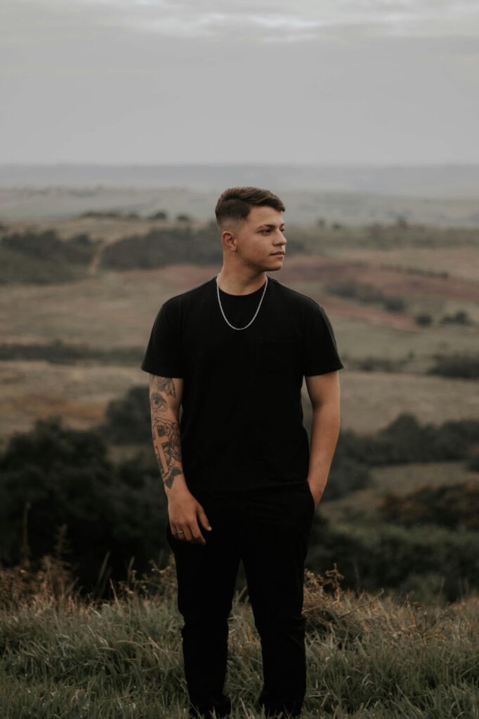 Man standing outdoors in a black shirt and chain, overlooking a serene green countryside with a calm and reflective expression.