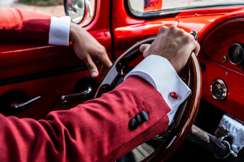 A man in a vibrant red suit drives a vintage car with a classic wooden steering wheel.