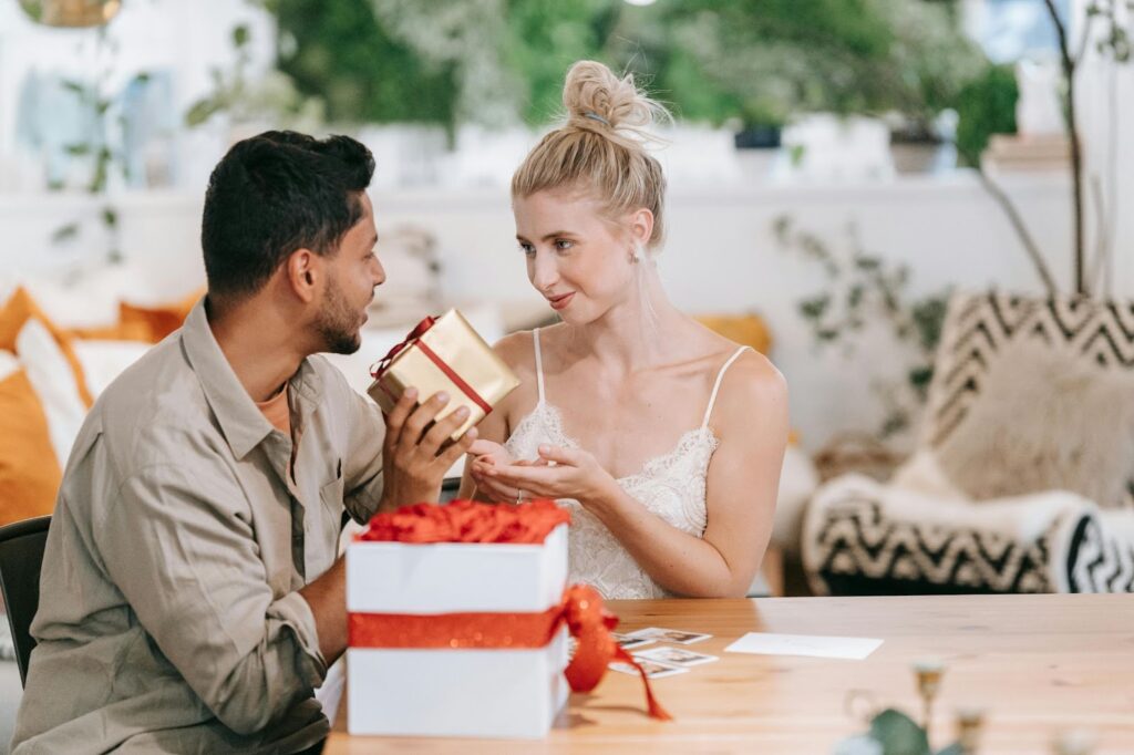 Couple exchanging a gift in a cozy setting, with a gold-wrapped present and a festive box on a wooden table, surrounded by warm decor.