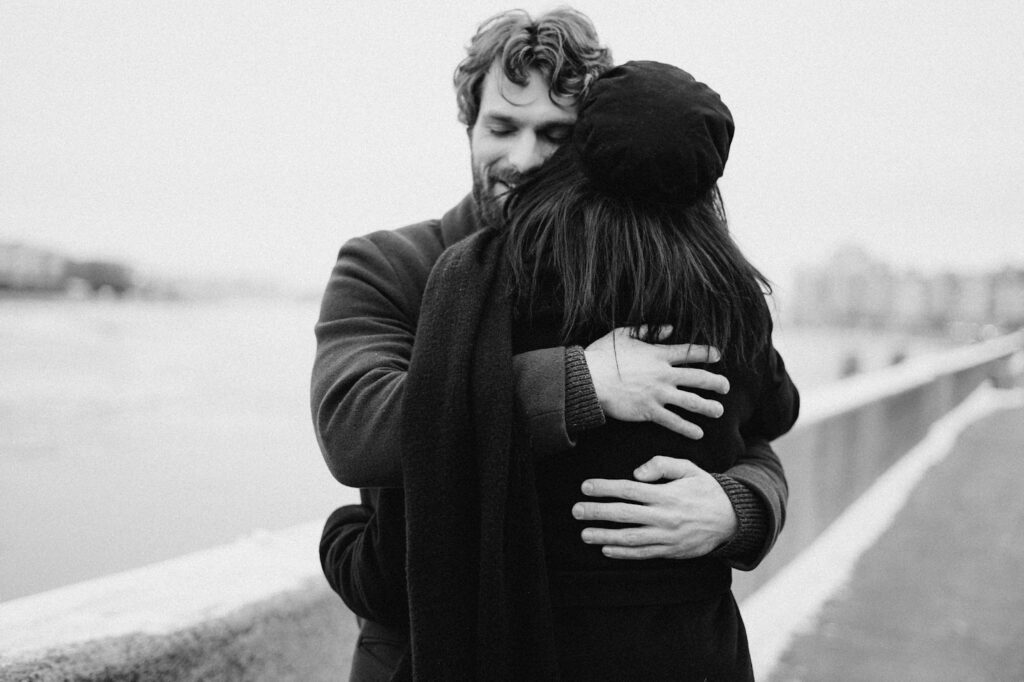 Black and white image of a couple embracing warmly on a riverside walkway, with soft expressions and a peaceful background view.