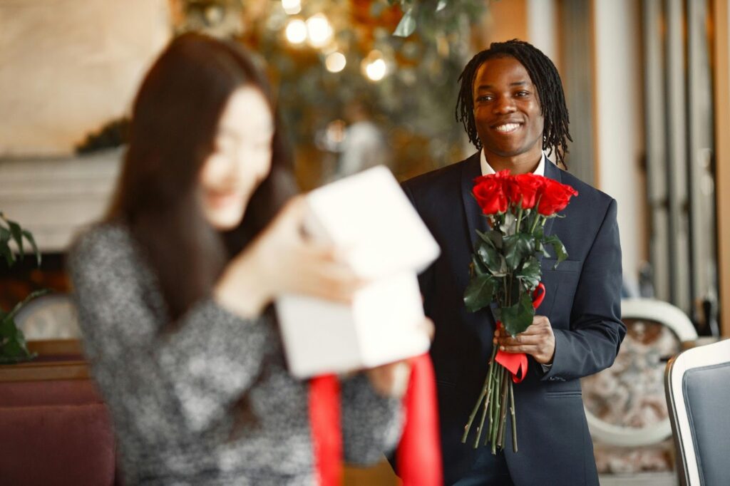 A man in a navy suit holds a bouquet of red roses, smiling warmly as a woman excitedly opens a gift in a cozy, softly lit setting.