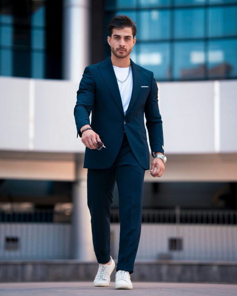 A stylish man in a navy suit, white shirt, and sneakers confidently walks outdoors, holding sunglasses, with a modern building in the background.