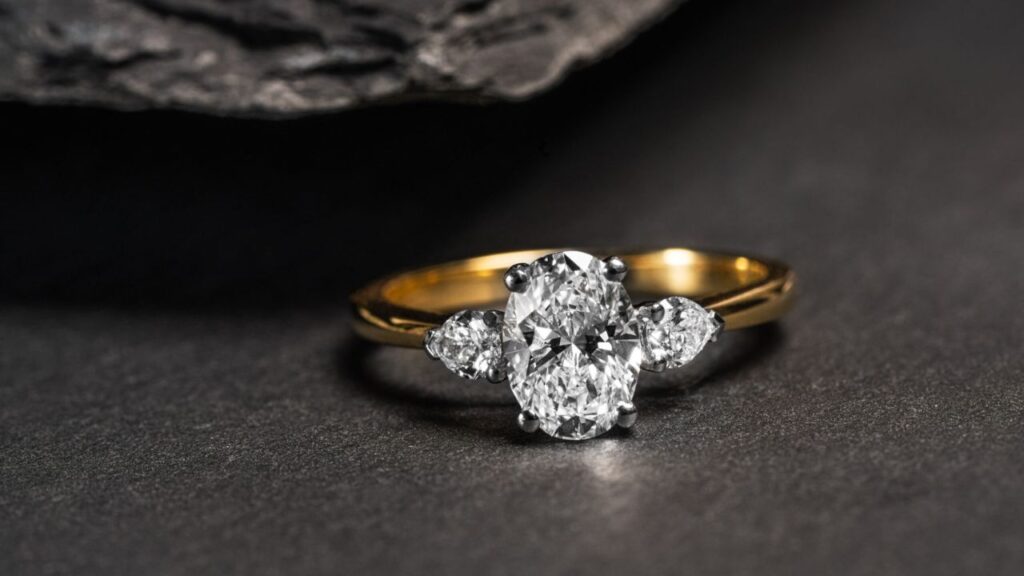 A gold ring featuring an oval diamond flanked by two smaller diamonds, resting on a dark, textured surface.