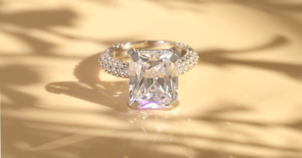 A large, emerald-cut diamond ring with a pave diamond band, resting on a reflective surface with warm, golden lighting.