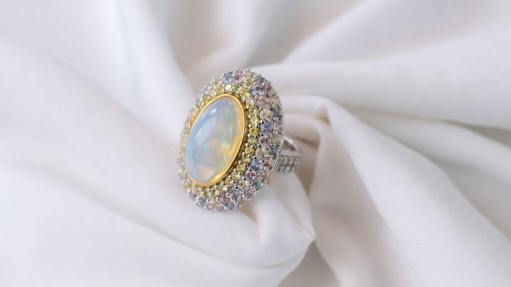 A large opal ring surrounded by smaller diamonds and multi-colored sapphires, resting on a soft, white, draped fabric.