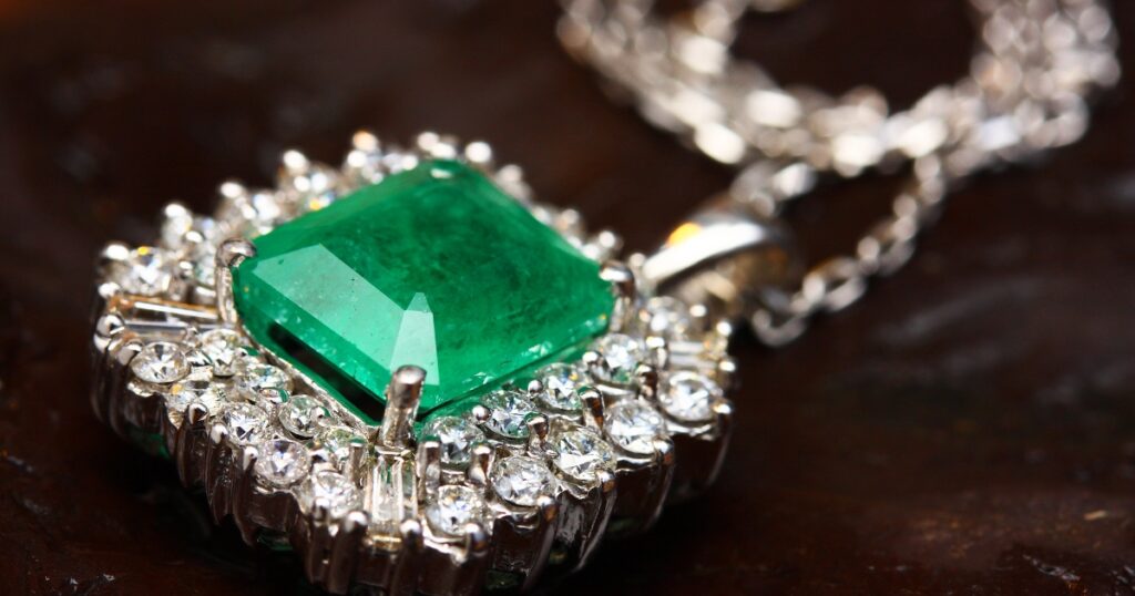 A close-up of a silver necklace featuring a large, square-cut emerald pendant surrounded by a halo of diamonds.